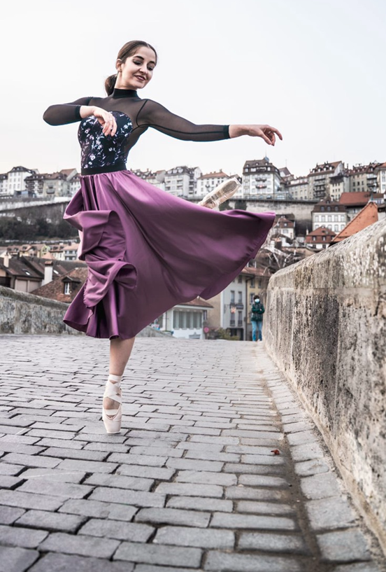 In this image, Katja is wearing a black top with a purple skirt and ballet shows. She has one leg in the air and her top half of her body is turned to face behind her. She is dancing on a stone bridge.