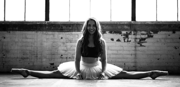 In this image, Katja is wearing a black crop top and a white ballet skirt, along with ballet shoes. She is doing the splits. The image has a black and white effect on it.